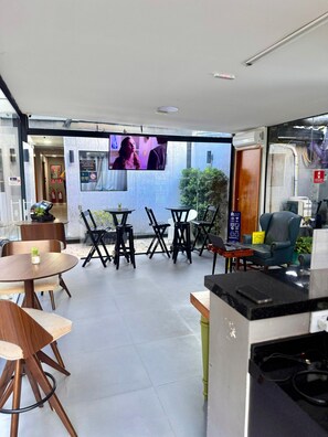 A modern interior with a flat-screen TV mounted on the wall, wooden tables and chairs, and a seating area with a sofa and armchairs. - ADD HOTÉIS MORUMBI (São Paulo)
