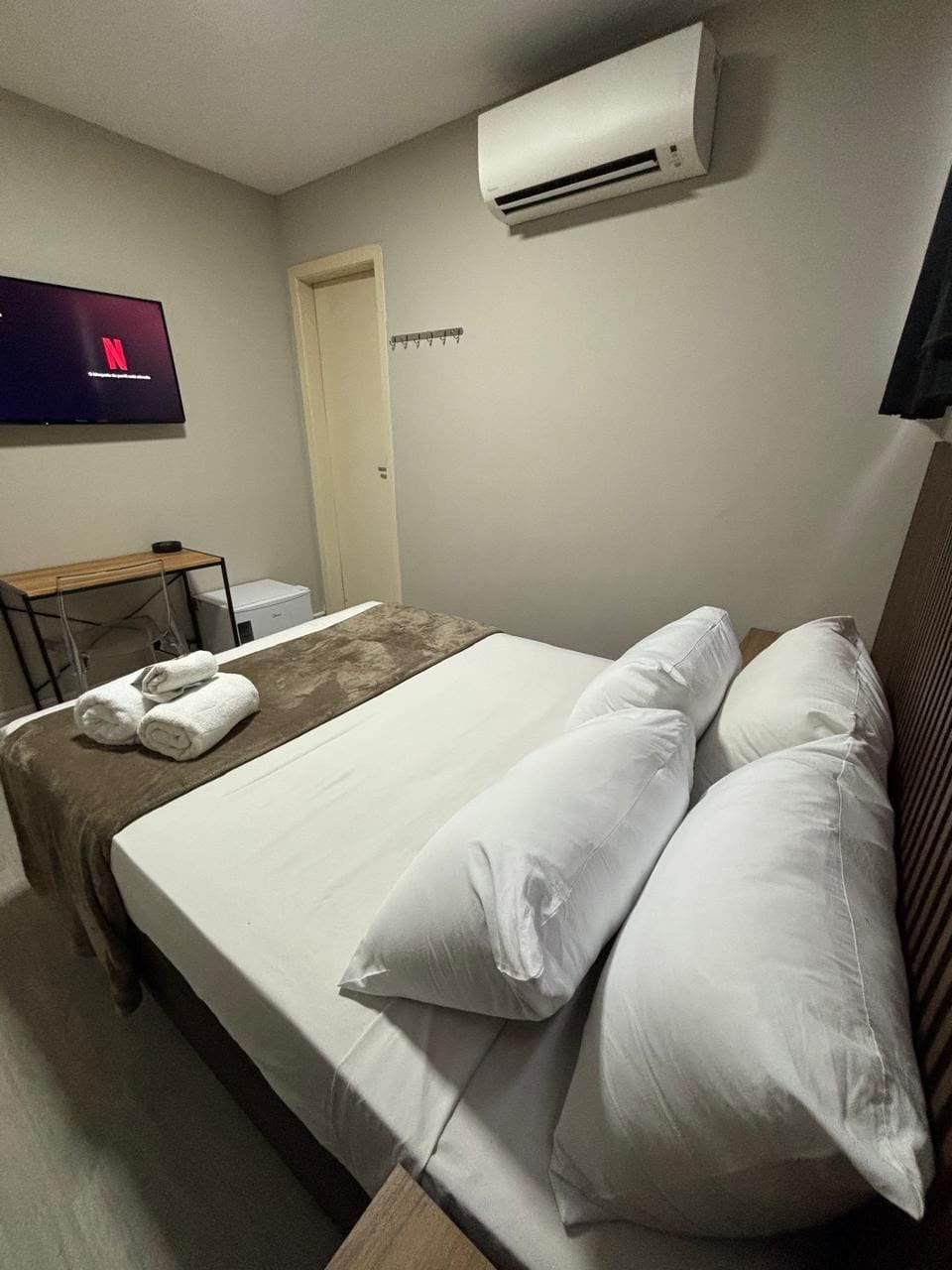 A hotel room with a bed, pillows, a TV, an air conditioner, and a desk with a chair.