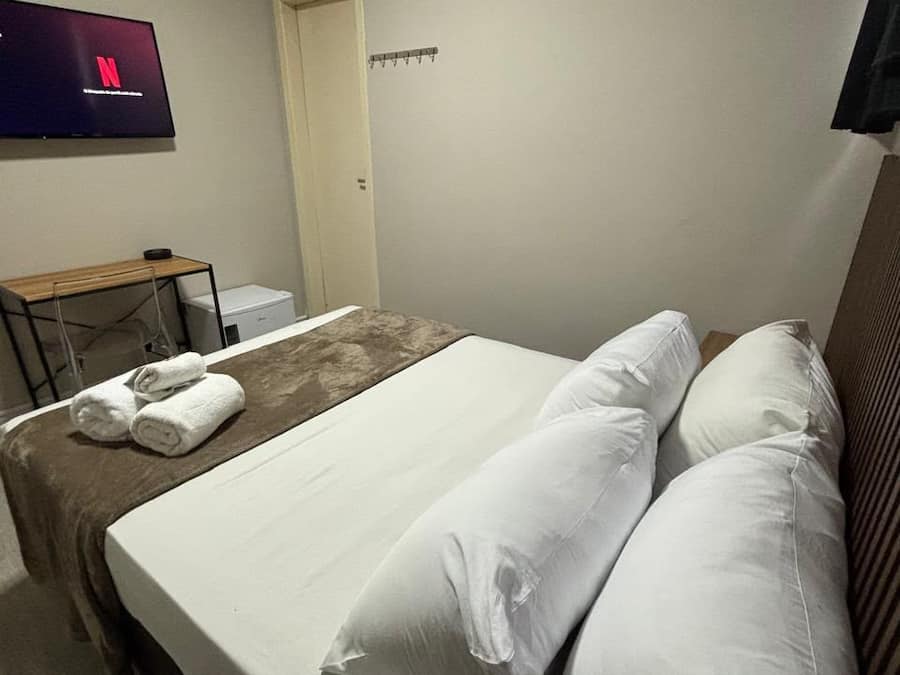 Quarto de hotel com cama, travesseiros, televisão, ar condicionado e escrivaninha com cadeira.