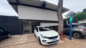 Un coche blanco aparcado frente a un edificio con una puerta de garaje negra, un árbol y un poste de seguridad azul.