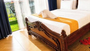 A hotel room with a wooden bed, a wooden nightstand, and a view of a green outdoor area.