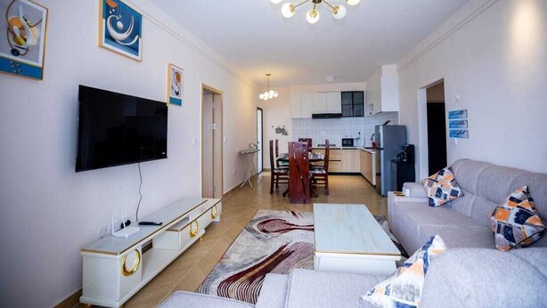 A modern living room with a flat-screen TV, a white entertainment unit, and a grey sofa with patterned cushions.