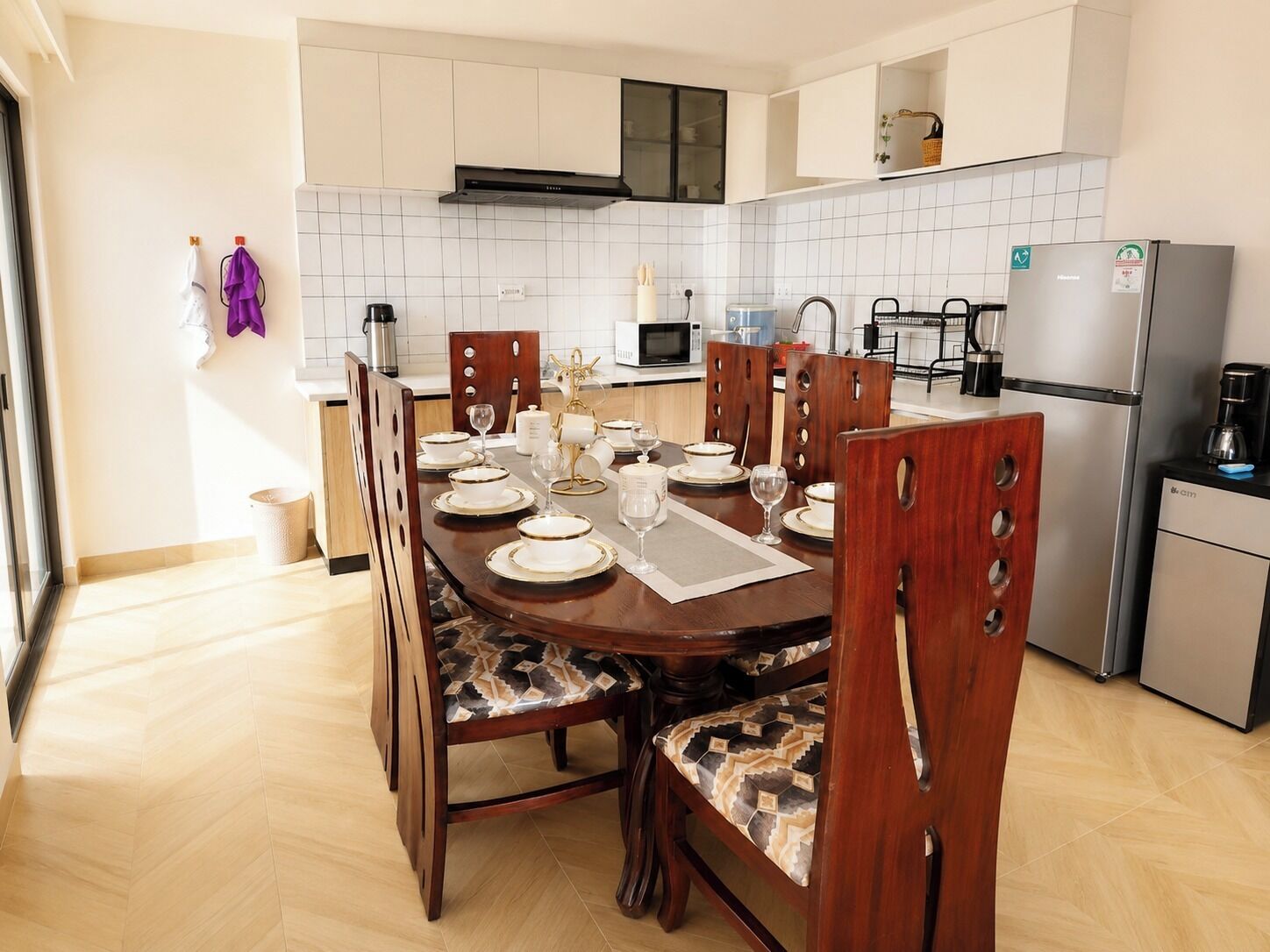 Una cocina moderna con armarios blancos, un refrigerador de acero inoxidable y un comedor para seis personas con sillas de madera y manteles estampados.