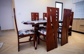 Un comedor con una mesa y sillas de madera, suelo de baldosas y una zona de cocina al fondo.