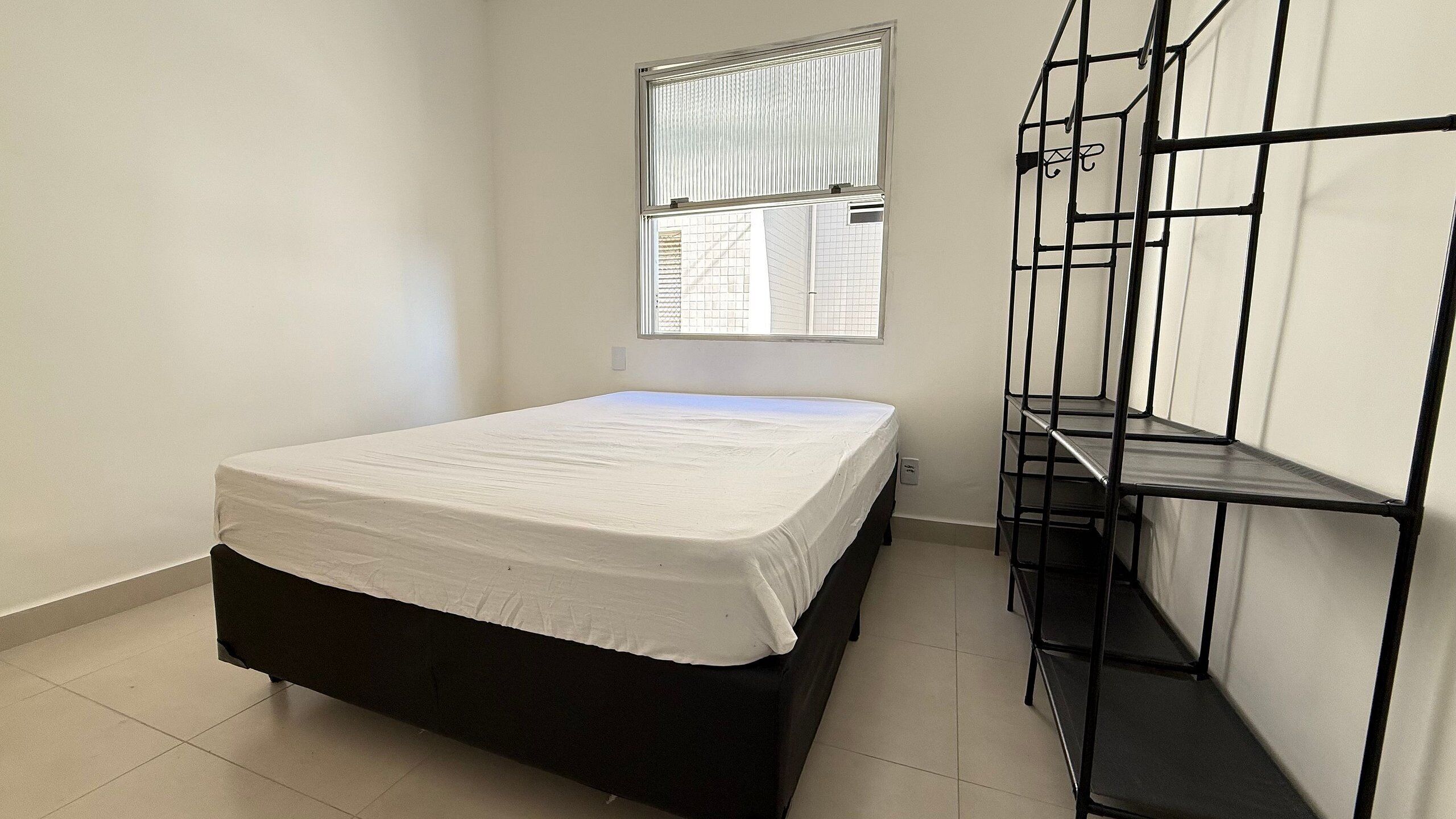 A room with a bed, a window with blinds, and a black metal shelf unit.