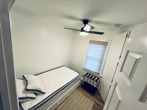 Ein kleiner, schmaler Raum mit einem Einzelbett, einem Deckenventilator, einem Fenster mit Jalousien und einer Tür.