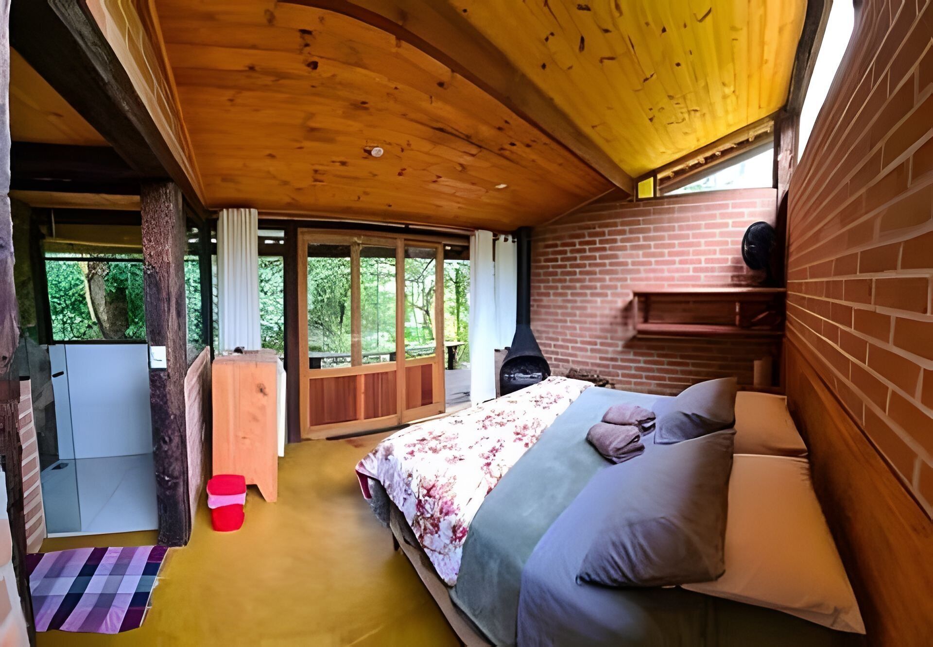 A bedroom with a wooden ceiling, a bed with floral bedding, a brick wall, and a fireplace.