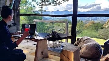 Una persona seduta a un tavolo di legno con un portatile e un bicchiere di bevanda rossa, con vista su un paesaggio montuoso attraverso una grande finestra.