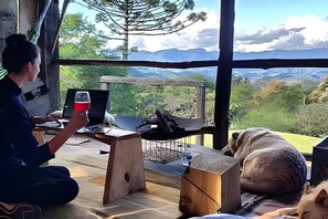 A person sitting at a wooden table with a laptop and a glass of red drink, overlooking a mountainous landscape through a large window. - Vida na Roça (São Bento do Sapucaí)