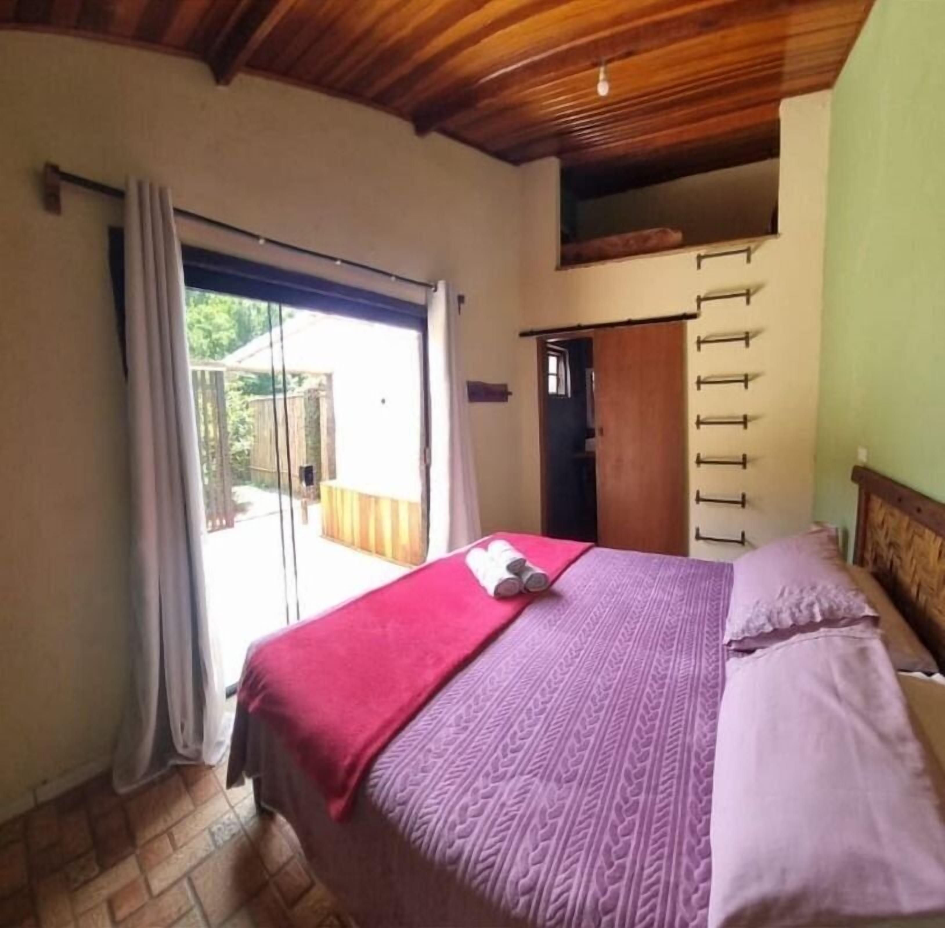 A bedroom with a bed, a window with curtains, a wooden ceiling, and a door leading to an outdoor area.