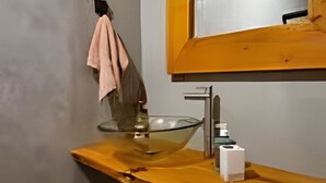 Un baño con tocador de madera, lavamanos de vidrio, una toalla colgada en la pared y un espejo con marco de madera.