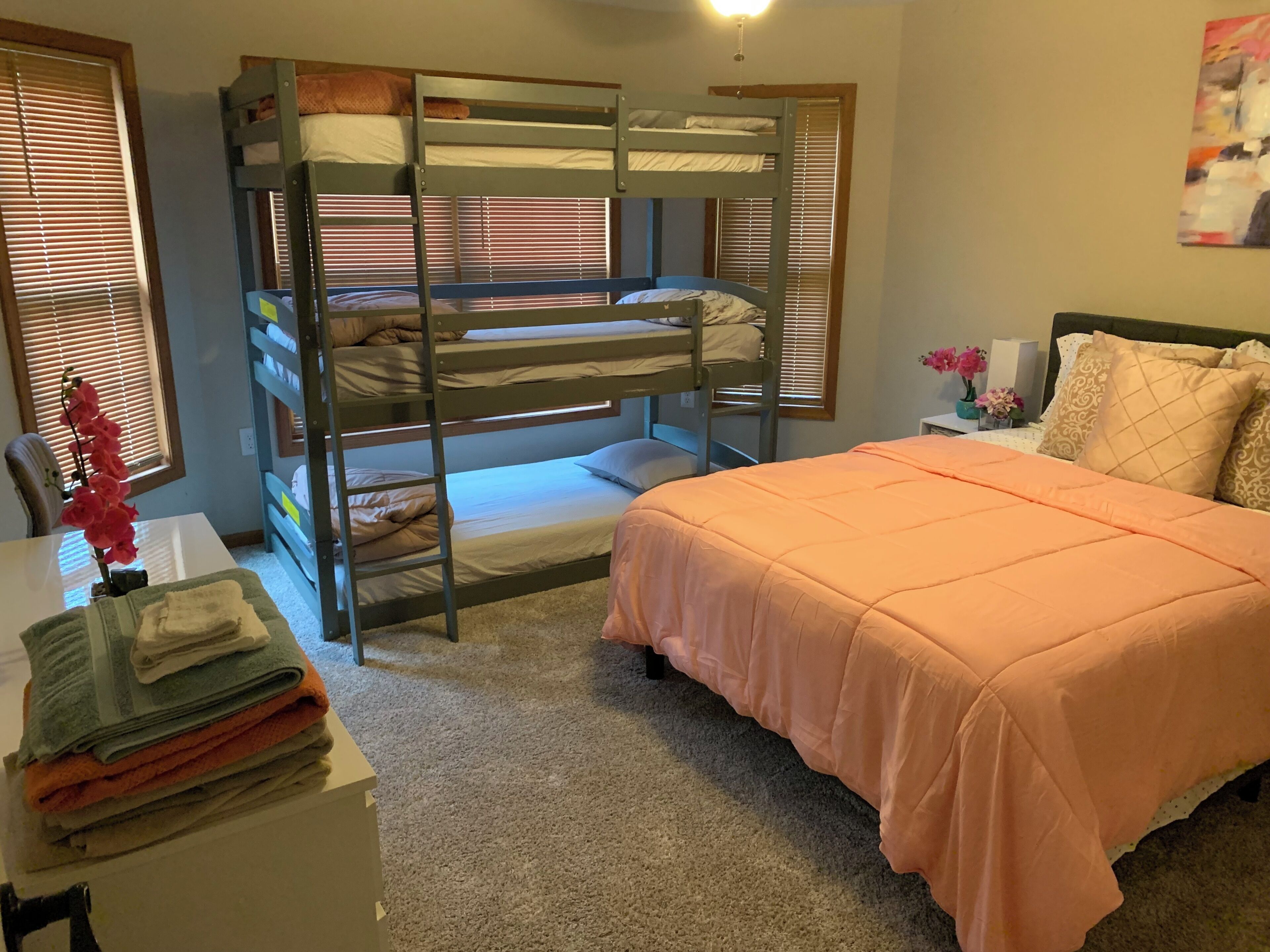 A bunk bed with a desk and chair, a bed with an orange bedspread, and a window with blinds.