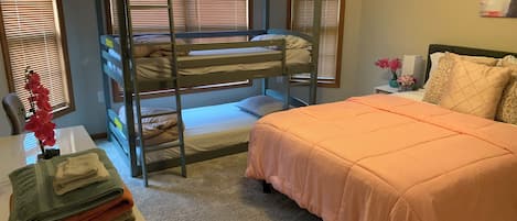 A bunk bed with a desk and chair, a bed with an orange bedspread, and a window with blinds.
