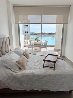 Ein Schlafzimmer mit Bett, Kissen, einem Nachttisch und Blick auf Pool und Gebäude durch ein großes Fenster.
