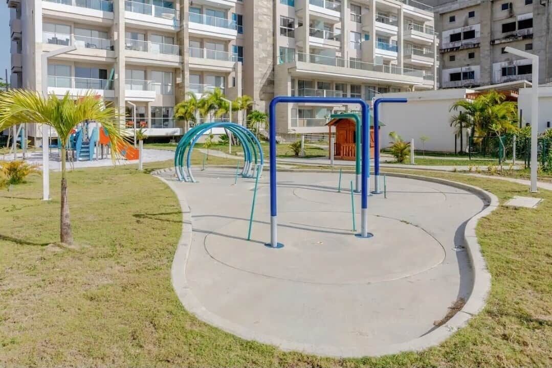An outdoor fitness area with exercise equipment, surrounded by a grassy area and palm trees, with a multi-story building in the background.