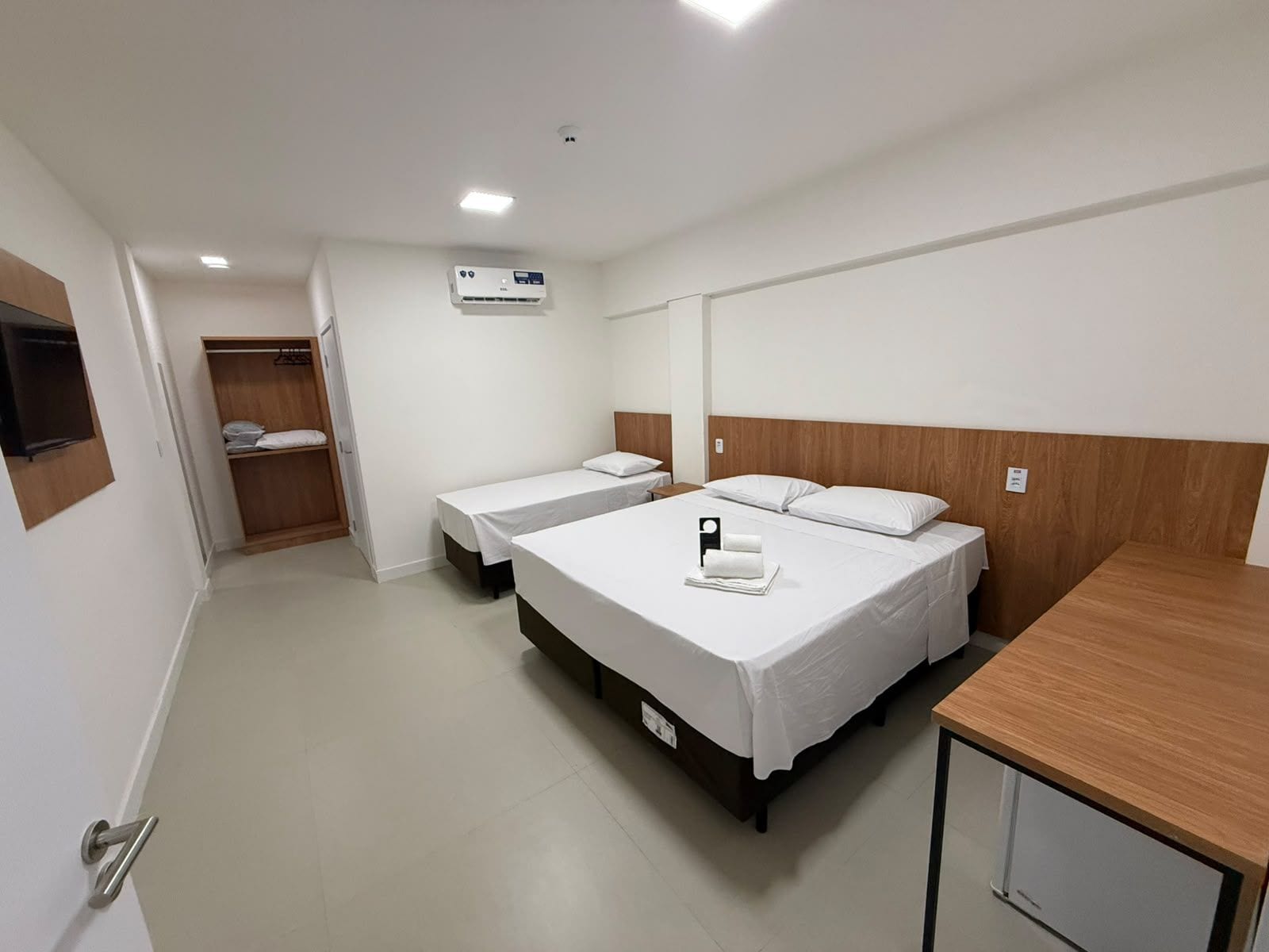 A hotel room with two single beds, a wooden headboard, a bedside table, a wardrobe, and a ceiling-mounted air conditioning unit.