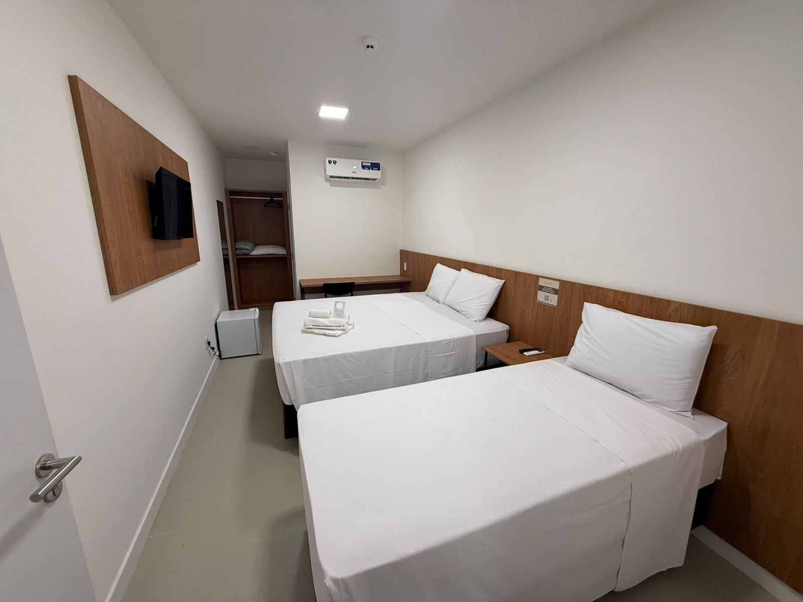 A hotel room with two beds, a wooden headboard, a bedside table, a small desk, a chair, a wardrobe, and a wall-mounted TV.