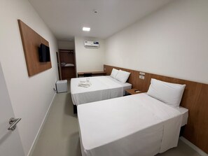 Quarto de hotel com duas camas, cabeceira de madeira, mesa de cabeceira, escrivaninha pequena, cadeira, guarda-roupa e televisão na parede.