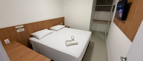 A hotel room with a bed, a wooden headboard, a small desk, a television mounted on the wall, and an air conditioning unit.