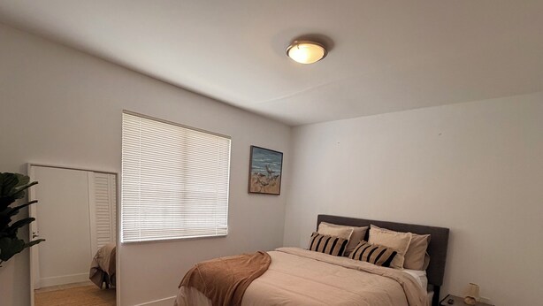 A bedroom with a bed, a bedside table, a mirror, a window with blinds, and a ceiling light. - Modern Home • Walk to Design Dist• Free Parking (Miami)