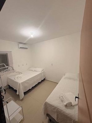 A clean, well-lit room with two beds, a window with blinds, an air conditioning unit, and a white chair.