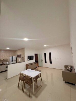 A spacious living area with a dining table and chairs, a sofa, a kitchen area with appliances, and a television mounted on the wall.