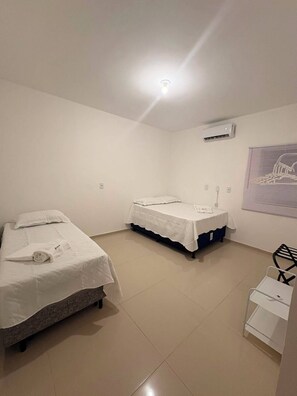 Quarto de hotel com duas camas, uma parede branca, um ar condicionado, uma janela com persianas e uma pequena mesa branca.