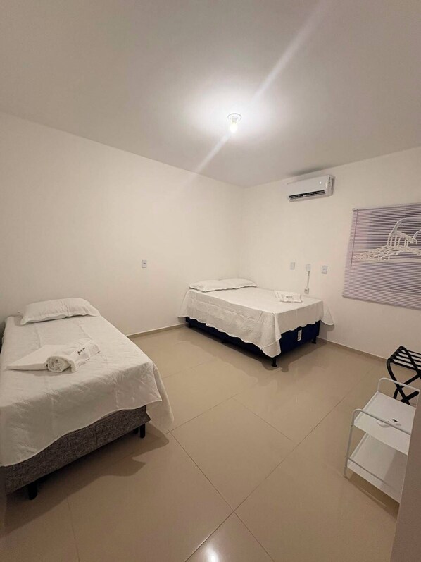 A hotel room with two beds, a white wall, an air conditioning unit, a window with blinds, and a small white table.