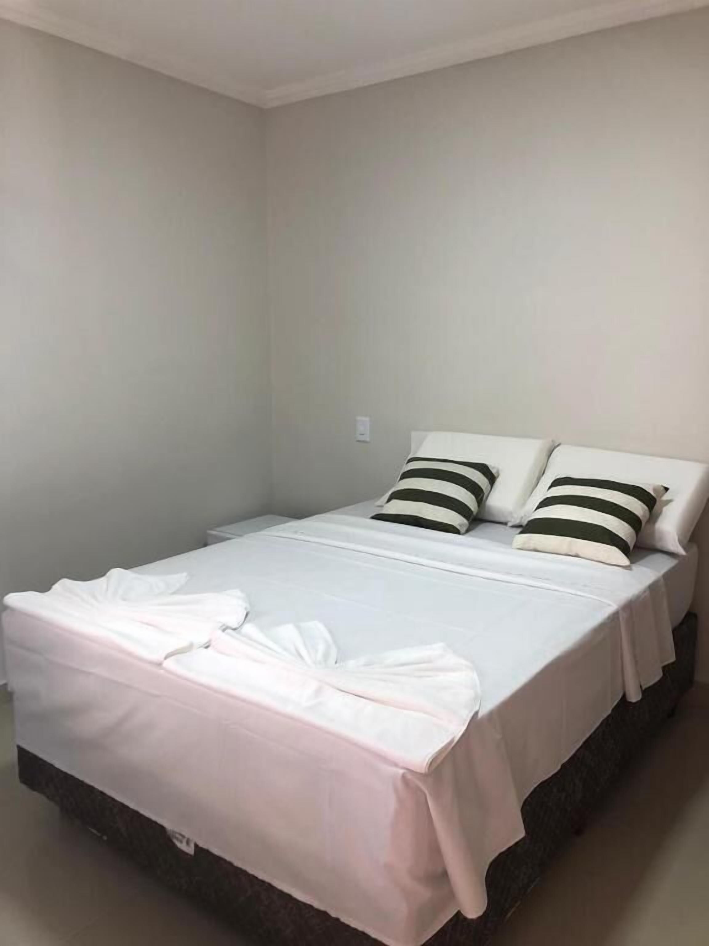 A single bed with white bedding and striped pillows, a plain wall, and a small light switch.
