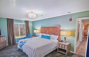 Una habitación de hotel con cama, mesitas de noche, ventana con cortinas y puerta que da a otra habitación.
