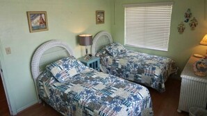 Una camera da letto con due letti, un ventilatore a soffitto, una finestra con persiane e un tavolino con una lampada.