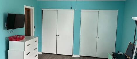 Una habitación con paredes verde azulado, un ventilador de techo, un aparador blanco con cajones, una cesta roja, un televisor negro, una puerta blanca y una mesa negra con una silla.