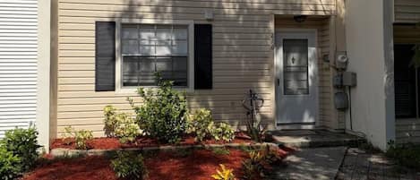 A two-story building with a front door, windows, and a small garden with red mulch and plants.