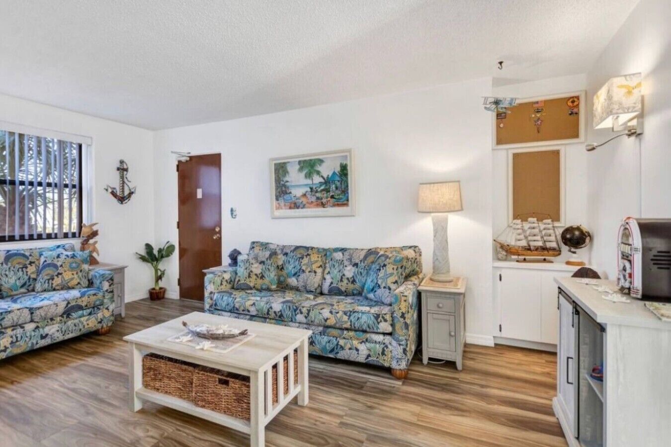A living room with two sofas, a coffee table, a lamp, and a kitchen area with cabinets and a refrigerator.
