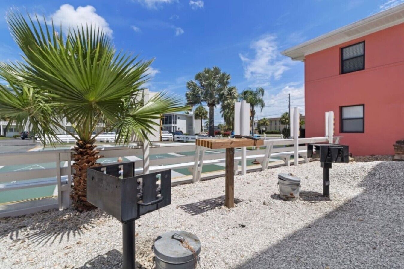 A parking area with a palm tree, a red building, and a tennis court in the background.