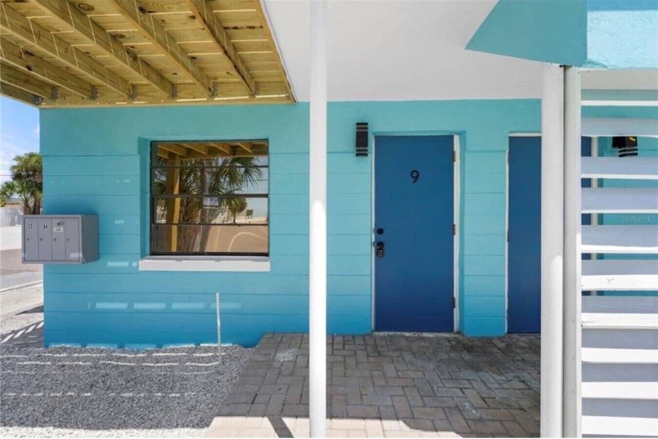 A building with a blue exterior, a door marked with the number 9, and a window with a view of palm trees.