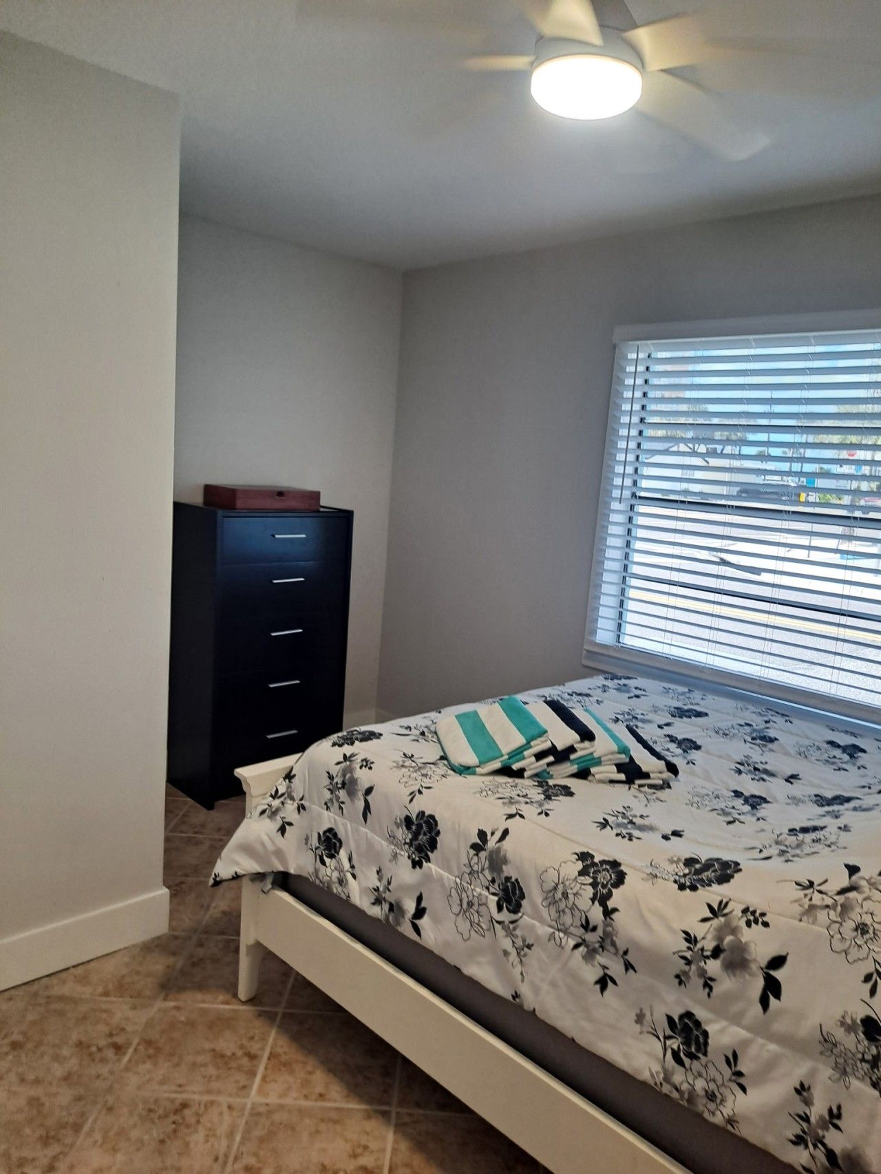 Un dormitorio con cama, cómoda, ventilador de techo y ventana con persianas.