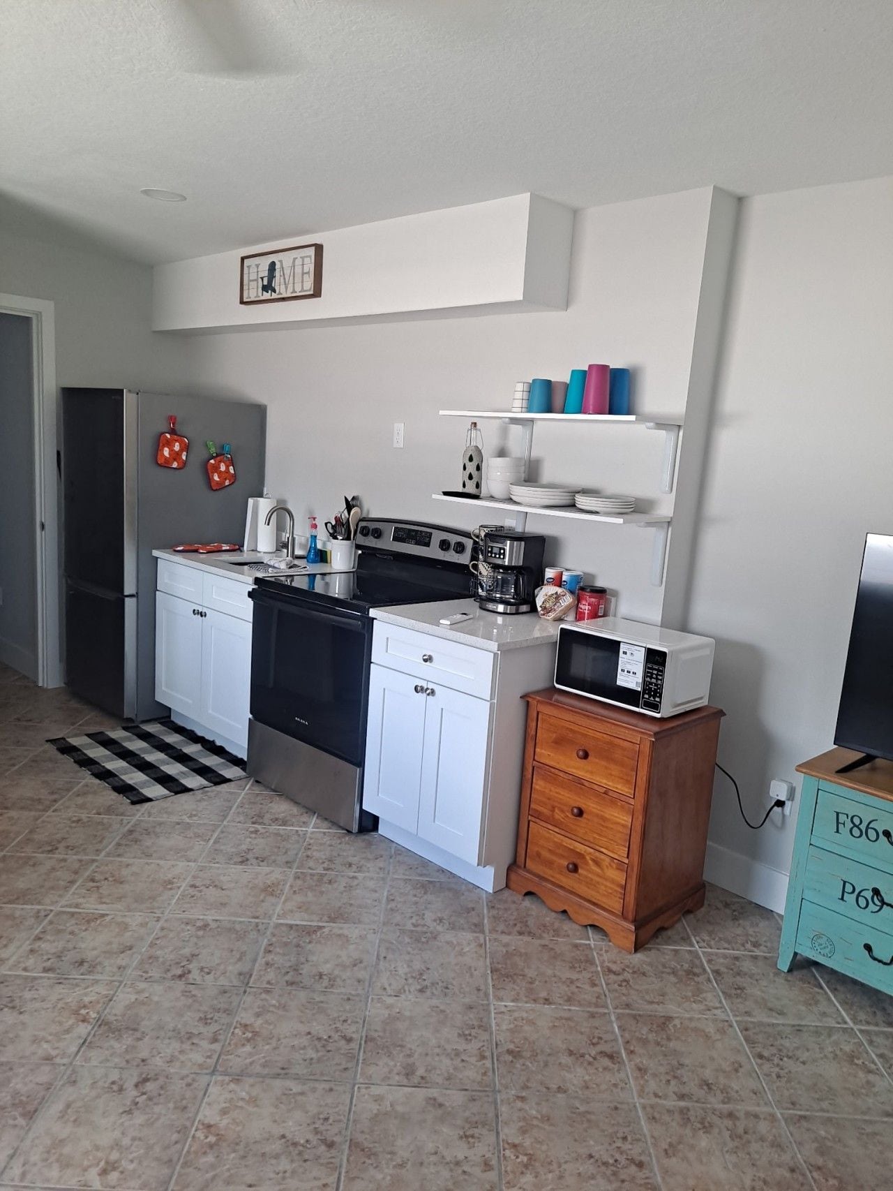 Cozinha compacta com armários brancos, geladeira de aço inoxidável, fogão e micro-ondas. Há um armário de madeira com gavetas e uma pequena prateleira com xícaras.