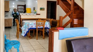 A dining area with a table set for four, a wooden staircase, and a kitchen in the background. - Casa Estrela Cond Fechado c Piscina - RJ (Cabo Frio)