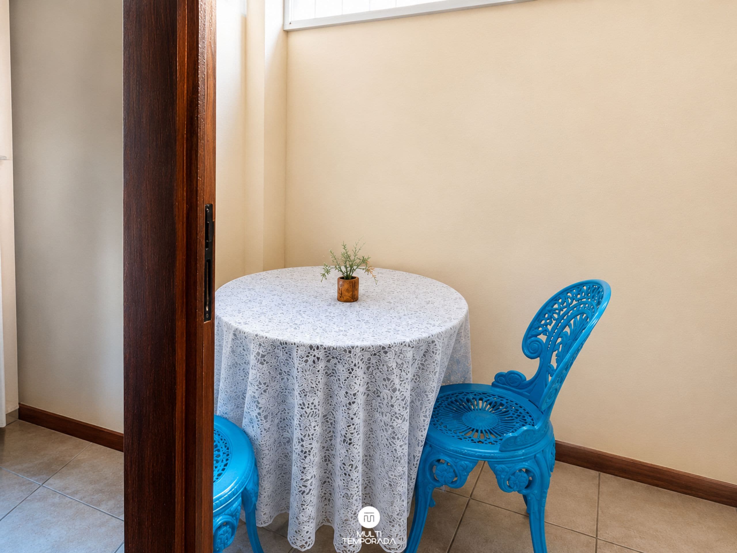 A small round table with a lace tablecloth, a potted plant, and two blue chairs in a corner of a room with beige walls and a wooden floor.