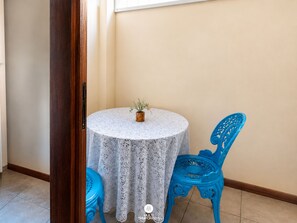 Une petite table ronde recouverte d’une nappe en dentelle, un pot de fleurs et deux chaises bleues, le tout placé dans un coin d’une pièce aux murs de couleur beige et au plancher en bois.