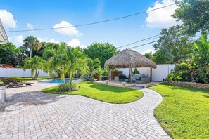 Ein Garten mit Swimmingpool, einem Reetdachgebäude und einer gepflasterten Terrasse.
