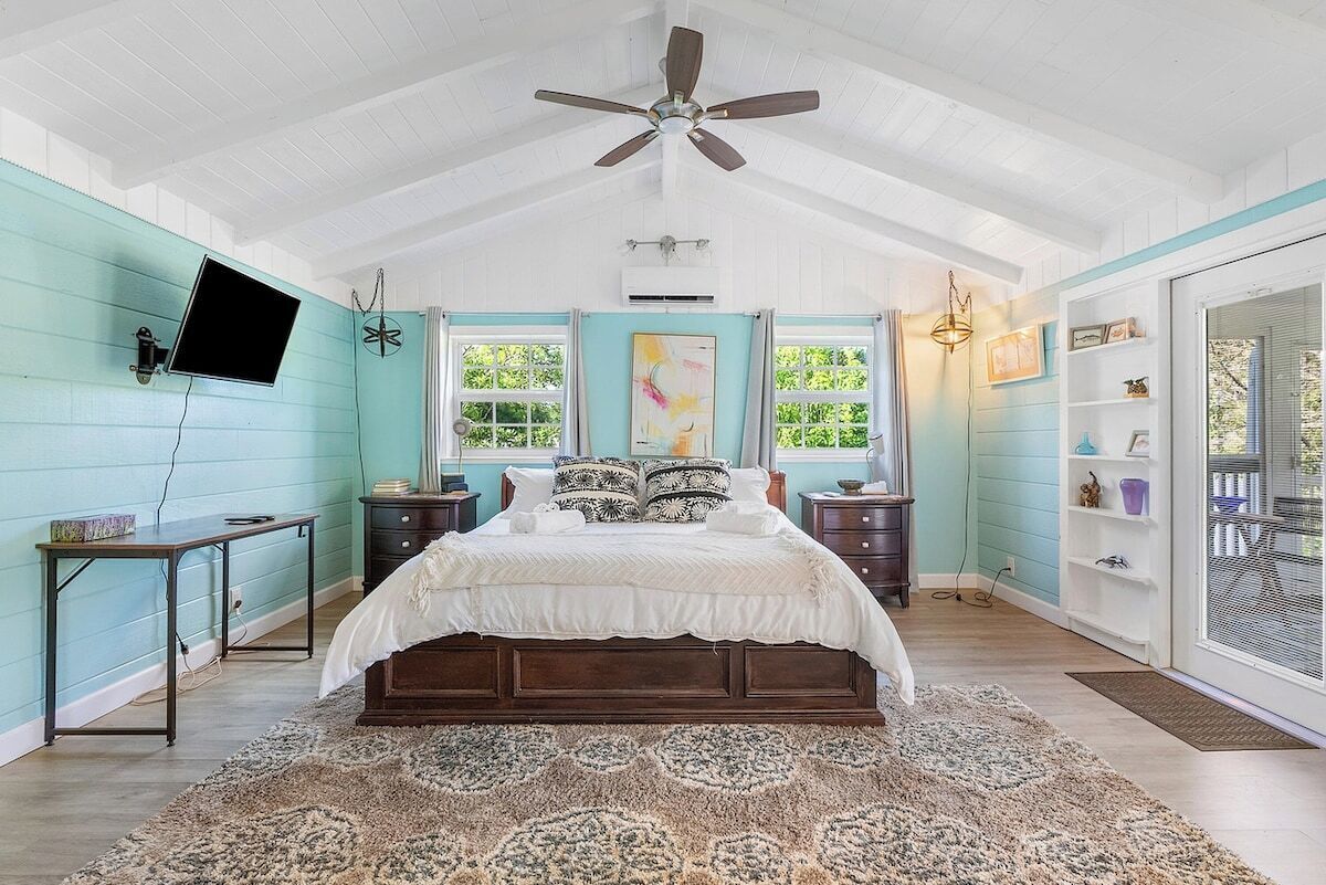 A bedroom with a bed, a ceiling fan, a TV mounted on the wall, and a window with curtains.