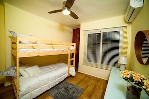 Ein kompaktes Zimmer mit Etagenbett, Deckenventilator, Klimaanlage, Fenster mit Jalousien sowie einem kleinen Tisch mit Lampe und Blumen.