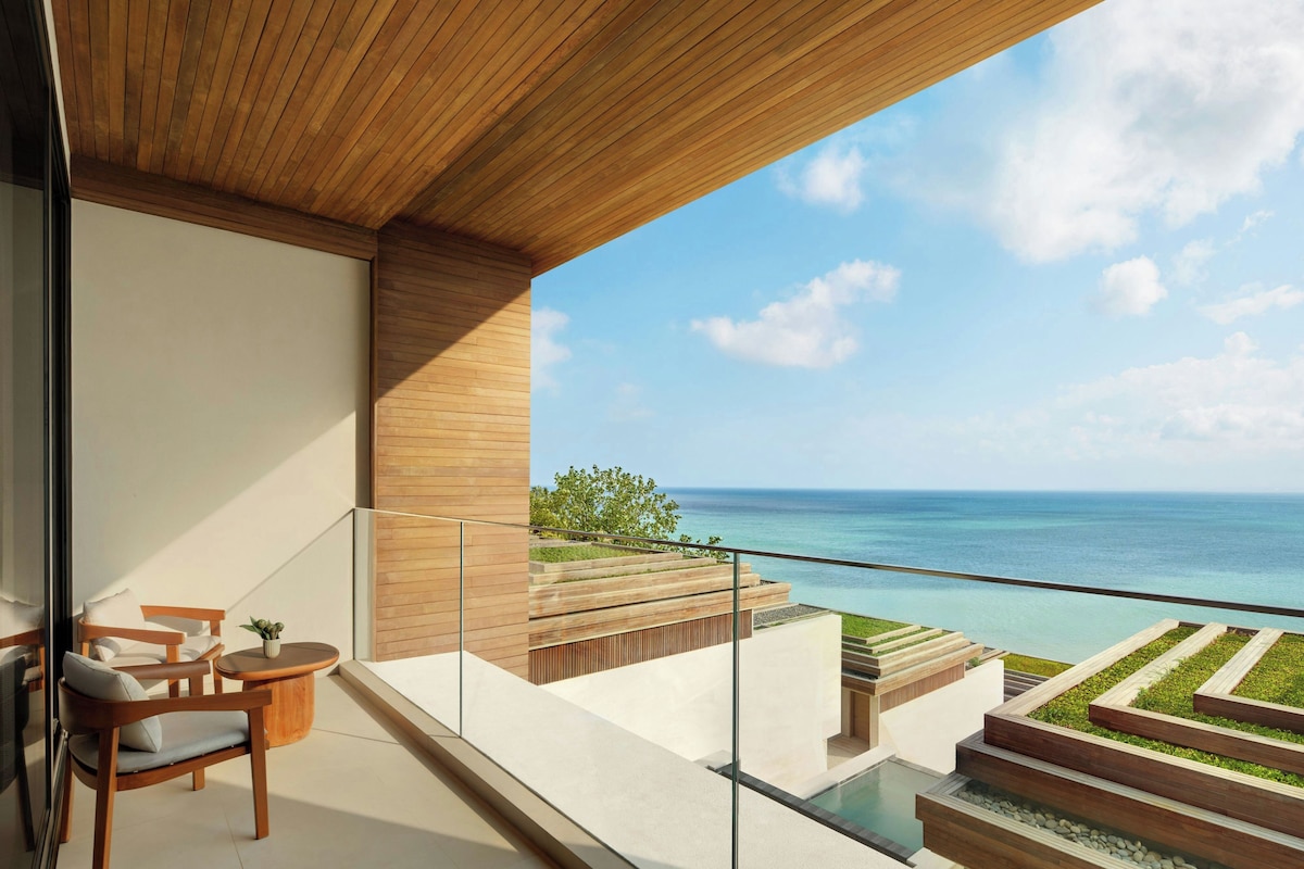 A modern balcony with a view of the ocean, wooden furniture, and a pool area.