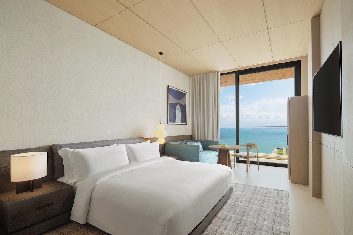 A hotel room with a large bed, a desk, a chair, a TV, and a view of the sea.