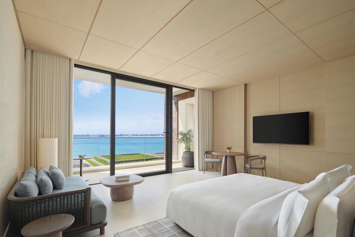 A modern hotel room with a large bed, a flat-screen TV, a seating area with a view of the sea, and a balcony.