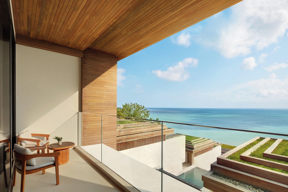 A modern balcony with a wooden ceiling, a seating area with a chair and table, and a view of the ocean.