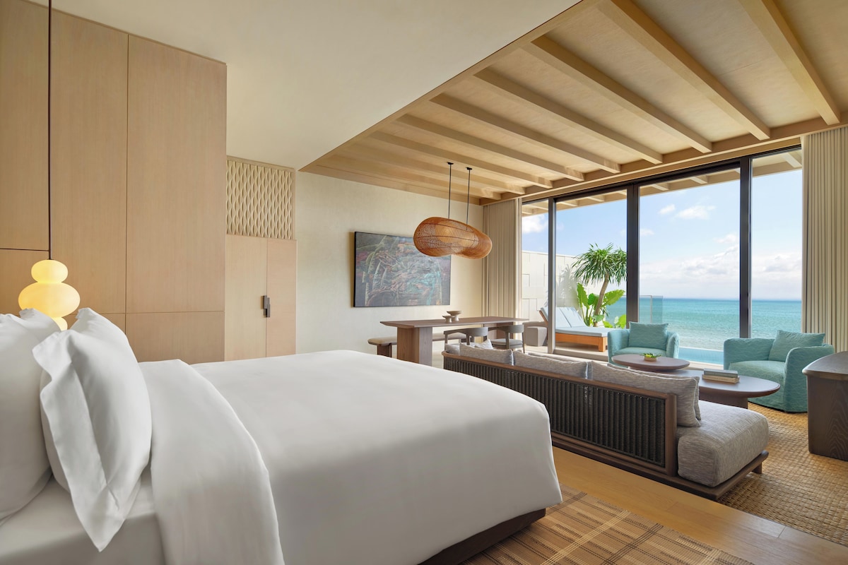 A modern hotel room with a large bed, a dining area, and a view of the ocean.