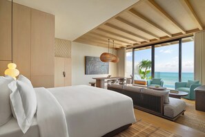 A modern hotel room with a large bed, a dining area, and a view of the ocean.
